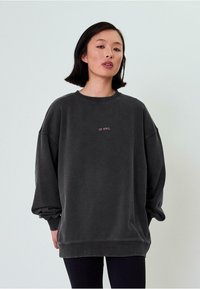 Übergroßer grauer Sweatshirt mit Rundhalsausschnitt, langen Ärmeln und pinkfarbenem gesticktem Text auf der Vorderseite. Weicher, lockerer Schnitt, gerippte Bündchen.