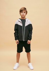 Svart och grå lättviktsjacka med dragkedja, tillsammans med svarta shorts och vita sneakers på en neutral bakgrund.