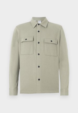 Ljust beige långärmad skjortjacka med krage, svarta tryckknappar och två framfickor med klaff, uppvisad mot en vit bakgrund.