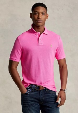 Giovane in camicia polo rosa brillante e jeans scuri, con una cintura a righe, in piedi con sicurezza di fronte a uno sfondo neutro.
