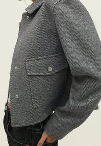 Szary żakiet w wzór herringbone z kołnierzykiem, zatrzaskami i boczną kieszenią, o dopasowanym kroju i teksturowanej wełnianej tkaninie.