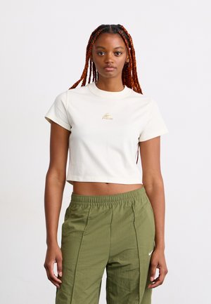 Young woman with long red braids wearing a white cropped t-shirt and olive green pants standing against a plain white background.