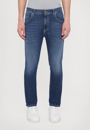 Homme portant un jean bleu slim fit et des baskets blanches, debout devant un fond clair uni.