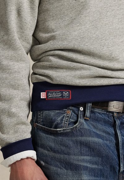 Grey sweatshirt with navy ribbed hem, featuring a red label with white text. Worn with faded blue denim jeans showing wear.