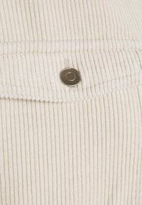 Detailní záběr béžové kordové látky s vertikálními pruhy a jedním kulatým stříbrným drukem na klopě kapsy.