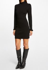 Robe noire à manches longues, silhouette ajustée, tissu texturé, col à volants et poignets à volants. Associée à de hautes bottes à talons en cuir noir.