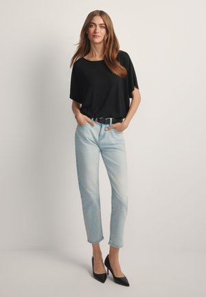 Kobieta stojąca z rękami w kieszeniach jasnoniebieskich dżinsów, ubrana w luźną czarną bluzkę i czarne buty na wysokim obcasie na jednolitym tle.