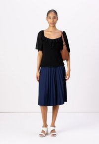 Schwarzes Oberteil mit Puffärmeln und Rüschendetails, kombiniert mit einem marineblauen Faltenrock, weißen Sandalen und einer braunen Umhängetasche.