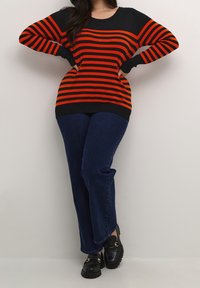 Pull en maille rayé rouge et noir à manches longues, associé à un jean droit bleu foncé et des mocassins noirs avec des accents dorés.