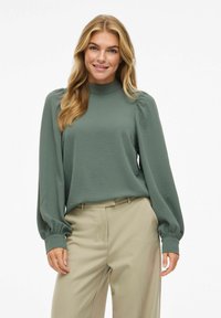 Blouse à manches longues, verte, avec un col haut, en tissu texturé et avec des poignets froncés. Associée à un pantalon beige.
