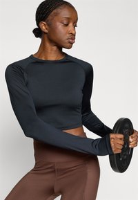 Juoda ilgomis rankovėmis trumpa palaidinė su raštuoto audinio detalėmis, derinama su aukštos juosmens rudos spalvos tamprėmis. Modelis laiko juodą svarmenų plokštę.