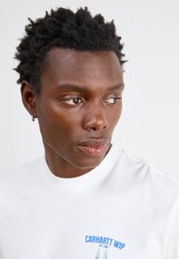T-shirt bianco in cotone con grafica blu "CARHARTT WIP" sul petto. Dettaglio ravvicinato di una persona con capelli neri corti e texture.