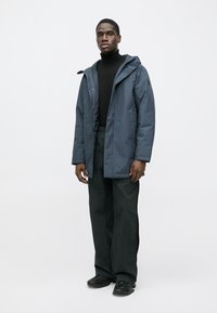 Σκούρο μπλε αδιάβροχο παρκά με ρυθμιζόμενο καπό, μπροστινά κουμπιά και κλείσιμο με φερμουάρ. Συνδυασμένο με μαύρο ζιβάγκο και φαρδιά, σκούρα πράσινα παντελόνια.