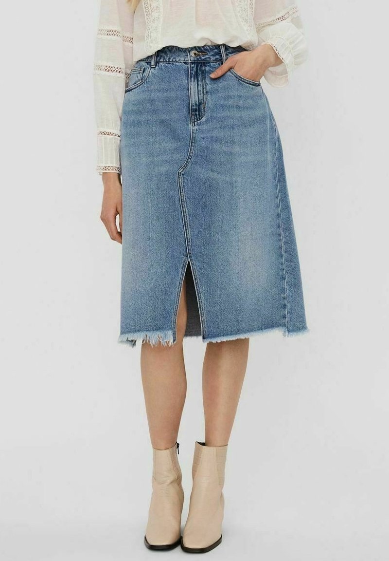 Femme portant une jupe en denim bleue longueur genou avec une fente avant et un ourlet brut, associée à des bottines beige et un chemisier blanc à manches longues.