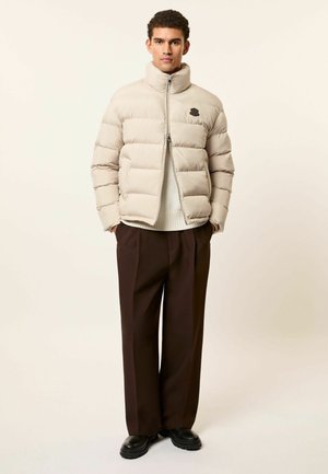 Mann steht, trägt eine beige Daunenjacke, weißen Pullover, braune Hose und schwarze Schuhe vor einem schlichten hellen Hintergrund.