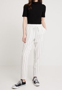 Kvinna iklädd svart kortärmad topp, vita byxor med snörning och tunna vertikala ränder, samt svarta låga sneakers som står med händerna i fickorna.