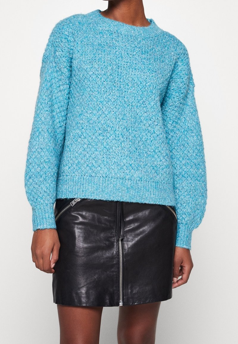 Pull en tricot bleu clair avec une finition texturée, col arrondi et manches volumineuses, accompagné d'une mini-jupe en cuir noir avec une fermeture éclair latérale.