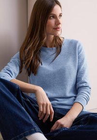  Lichtblauw, langemmouwen, textuurtrui met een ronde hals. Gecombineerd met hoge taille donkere denim, zittende pose tegen een neutrale achtergrond.