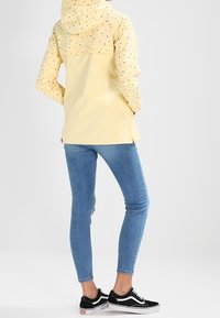 En person som bär en gul regnjacka med huva och små svarta mönster, blå tajta jeans och svartvita sneakers, står med händerna i fickorna.