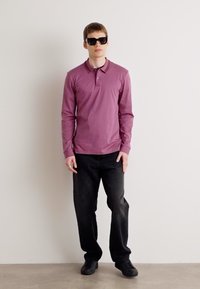 Camisa polo de manga longa, rosa clara, com gola, combinada com jeans pretos e sapatos pretos; modelo a usar óculos de sol escuros.