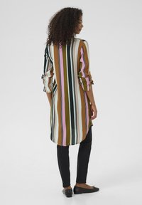 Chemise longue rayée dans des tons terreux de marron, vert, rose et blanc, avec une coupe décontractée, des manches retroussées et un ourlet incurvé, associée à un pantalon noir.