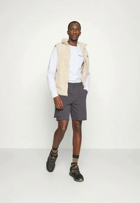 Άνδρας περπατάει φορώντας μπεζ φλις γιλέκο, λευκό πουκάμισο με μακριά μανίκια, σκούρες βερμούδες, ριγέ κάλτσες και μαύρα αθλητικά παπούτσια σε απλή φόντο.