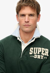 Groene katoenen rugbyshirt met een lichtblauwe binnenkraag, voorzien van een crèmekleurig logo en een twee-knopen placket. Zachte textuur, casual ontwerp.