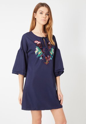 Joven mujer que lleva un vestido azul marino con mangas acampanadas y un diseño bordado colorido de pájaros y flores en el pecho.