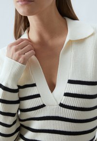Striped sweater in cream and black, featuring a v-neck and a ribbed texture. Long sleeves with cuff accents. Knitted fabric design.