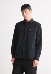 Donker corduroy overhemd met knopen, voorzien van een borstzak en een klein groen logo. Heeft een kraag en lange mouwen.