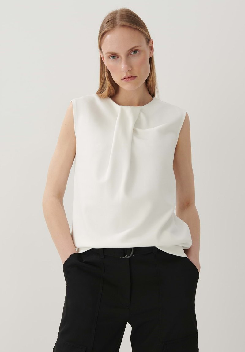 Weiße ärmellose Bluse mit einer Drapierung am Ausschnitt, kombiniert mit schwarzen Hosen. Glatter Stoff, minimalistisches Design.