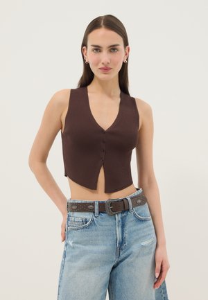 Mujer con un chaleco marrón acanalado sin mangas con botones y jeans de tiro alto azul claro con un cinturón marrón con tachuelas, de pie con una mano en la cadera.