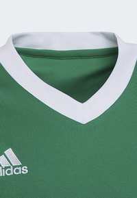 Jersey deportivo verde con cuello en V blanco, hecho de tela texturizada. Presenta un logo blanco bordado en el área del pecho izquierdo.