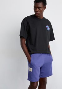 T-shirt en coton noir à manches courtes avec un graphique coloré sur la poitrine, associé à un short en polaire bleu-violet avec des poches latérales.