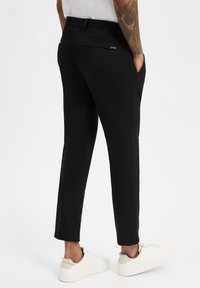 Schwarze, maßgeschneiderte Hose mit schmaler Passform, ausgestattet mit zwei Gesäßtaschen und einer glatten Stoffstruktur. Kombiniert mit weißen Sneakers.