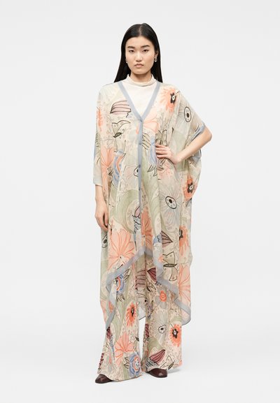 Γυναίκα στέκεται με το χέρι στη μέση, φορώντας διάφανο φλοράλ καμίαμο-στύλ κάρντιγκαν και ταιριαστά παντελόνια με φαρδιά μπατζάκια σε απαλά παστέλ χρώματα.