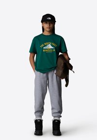 Green short-sleeve t-shirt with yellow print, gray sweatpants, black winter boots with fur trim, and a black cap, holding a brown bag.