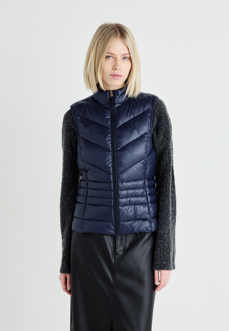 Navy puffer vest with chevron stitching, high collar, and zip front, paired with a dark gray long-sleeve sweater and a black leather skirt.
