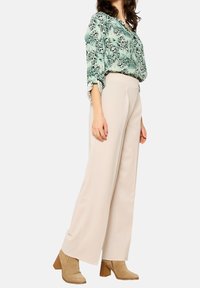 Svetle béžové kalhoty s širokým střihem a sklady, v kombinaci s blůzkou s zeleným vzorem. Modelka má na sobě hnědé vysoké boty na podpatku. Hladký materiál a volný střih.