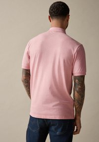 Camisa polo de manga corta rosa con cuello clásico, textura suave y corte entallado. Llevada con jeans azul oscuro, que presentan un detalle de brazo tatuado.