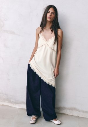 Giovane donna con lunghi capelli scuri che indossa una maglia crema con rifiniture in pizzo, pantaloni larghi blu navy e ballerine bianche, in piedi contro una parete chiara e testurizzata.