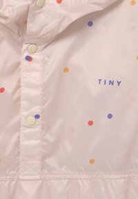 Tela de color rosa claro con lunares de colores en naranja, rojo y púrpura, botones de presión color crema y la palabra "TINY" impresa en púrpura.