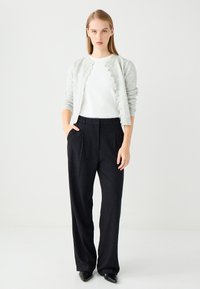 Femme debout les mains dans les poches, portant un cardigan boutonné gris clair, une chemise blanche, un pantalon noir à jambes larges et des chaussures pointues noires.