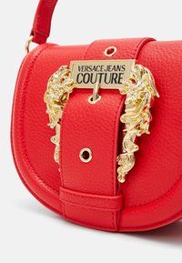 Punainen nahkainen olkalaukku, jossa on kultainen koristeellinen solki ja kaiverrettu "Versace Jeans Couture" -merkki etuylälaidassa.