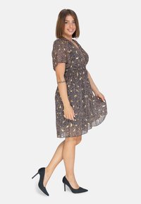 Vestido negro con flores, con mangas cortas abullonadas, cintura fruncida y dobladillo fluido. Lleva unos tacones de aguja negros. Fondo blanco.