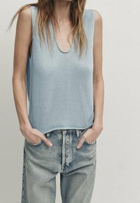 Vaaleansininen neulottu hihaton paita, jossa on löysä istuvuus, leveät hihansuut ja syvä pyöreä kaula-aukko, yhdistettynä korkeavyötäröisiin farkkuihin.