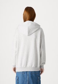 Siva prevelika dukserica s kapuljačom na vezanje, rebrastim manžetama i rubom. Izrađena od mekanog teksturiranog materijala, prikazana s leđa.