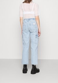 Žena v průhledném bílém krátkém topu, světle modrých vysokých cargo džínách a černých platformových kotníkových botách stojící zády k fotografu.