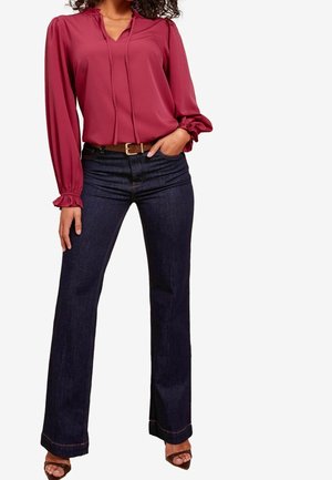 Vrouw draagt een lange mouwen bordeauxrode blouse met ruches aan de manchetten, donkerblauwe bootcut jeans, een bruine riem en open schoentjes, poserend met één hand in de zak.