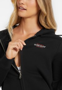 Crna hoodie na zip s kenguruzim džepom. Ima mali crveno-bijeli logo na prsima i kapuljaču sa vezicom. Mekan materijal.